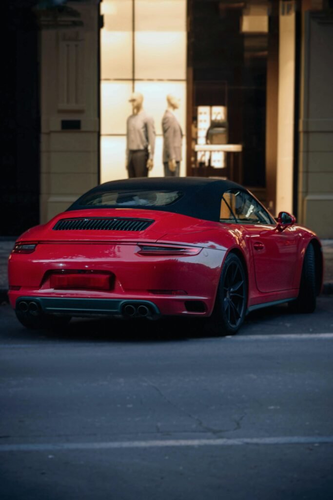 Sleek red sports car on a city street at dusk, with mannequins visible in a storefront.